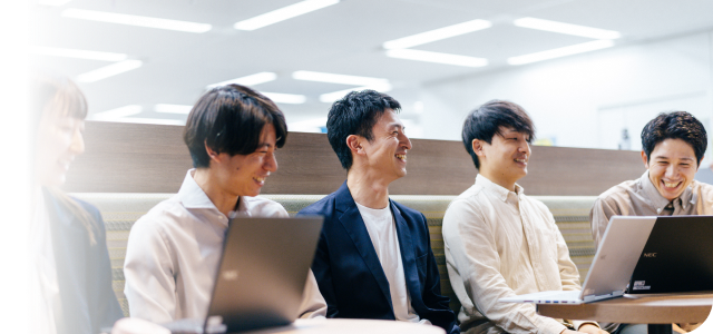 会議室で打ち合わせをしているビジネスパーソンたち。テーブルを囲んで5人の男女が和やかに会話をしながら、ラップトップや資料を活用して意見を交わしている。